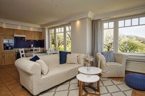 A spacious living room with a large beige sofa, a white coffee table, and a blue bean bag. A kitchen with wooden cabinets and a dining area with a white table and chairs is visible in the background.