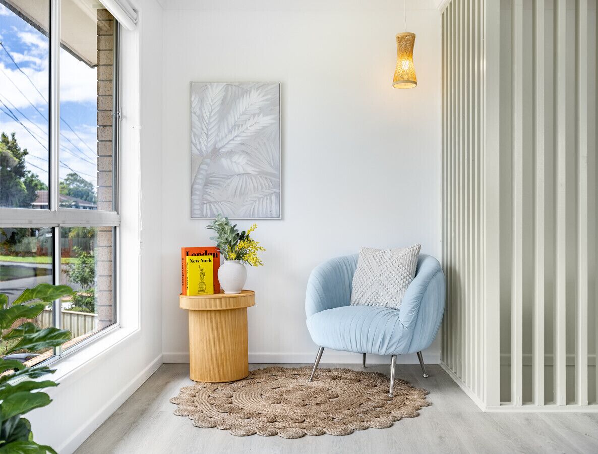 Une pièce d’angle avec une grande fenêtre, un fauteuil bleu, une petite table d’appoint en bois sur laquelle se trouvent un livre et des fleurs, un tapis tissé et un luminaire fixé au mur.