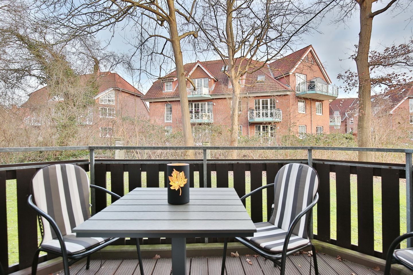 A balcony with a table and two chairs, overlooking a row of brick buildings with multiple windows and balconies.