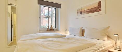 A bedroom with a large bed, a window with a view of buildings, a bedside table with a lamp, and a framed picture on the wall.