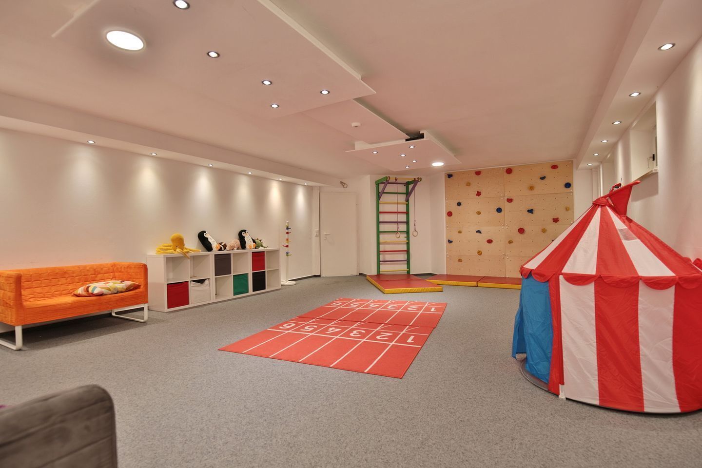 Una sala de juegos con una pared de escalar, una tienda roja y blanca y un juego de rayuela.