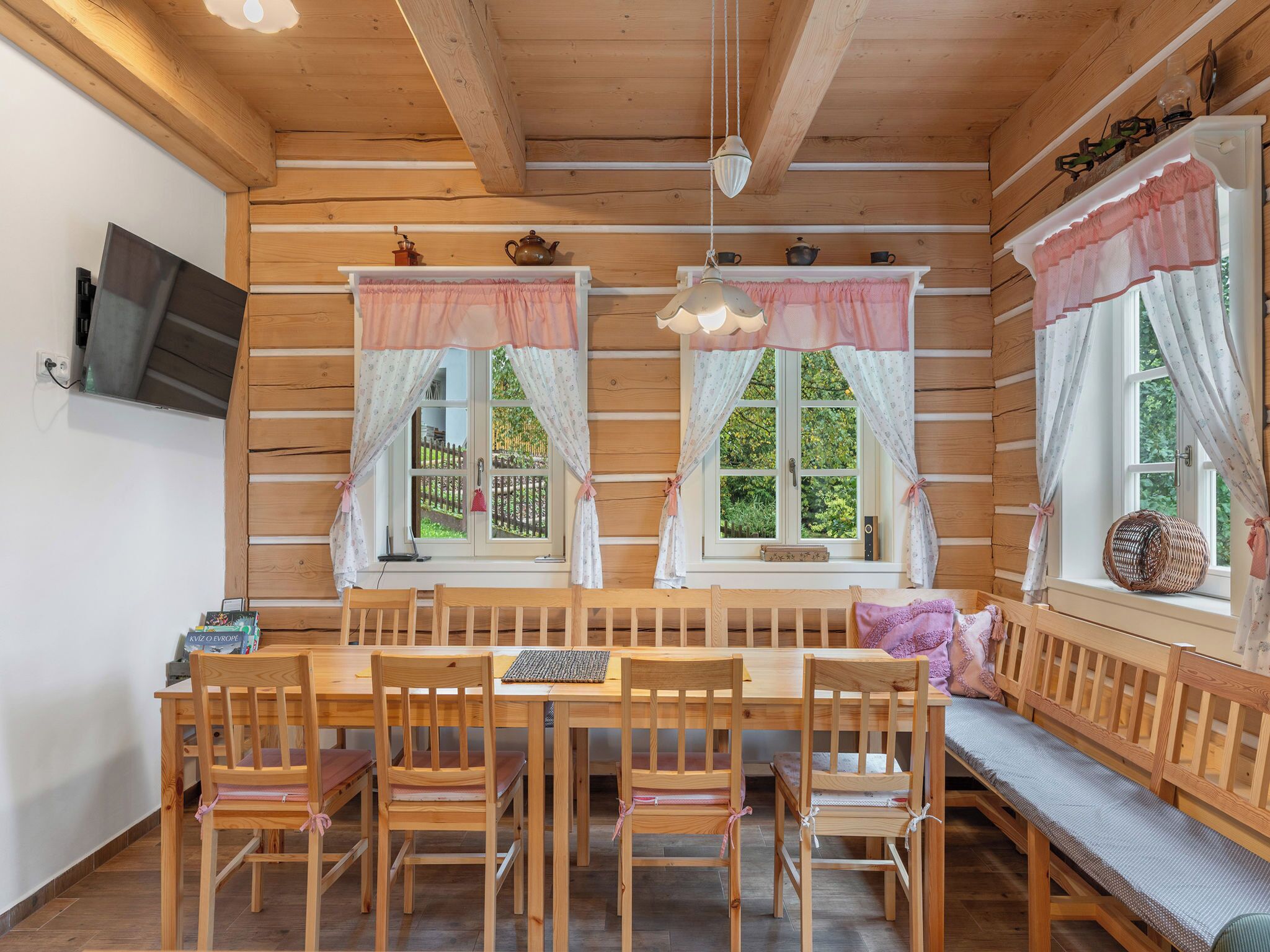 Una cabaña de madera con una mesa de comedor y sillas, dos ventanas con cortinas, y un televisor montado en la pared.