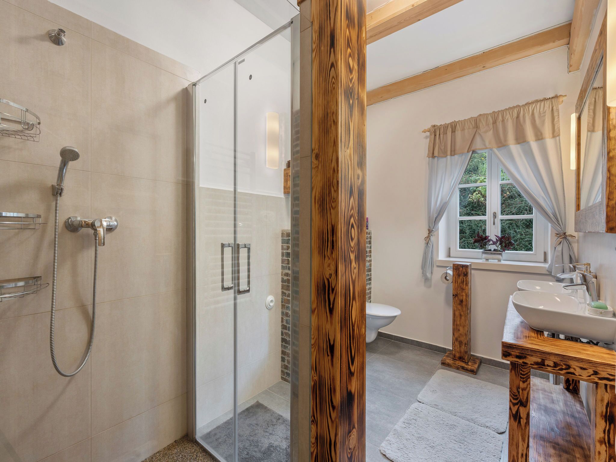Een moderne badkamer met een glazen douchecabine, een houten wastafelmeubel en een raam met gordijnen.