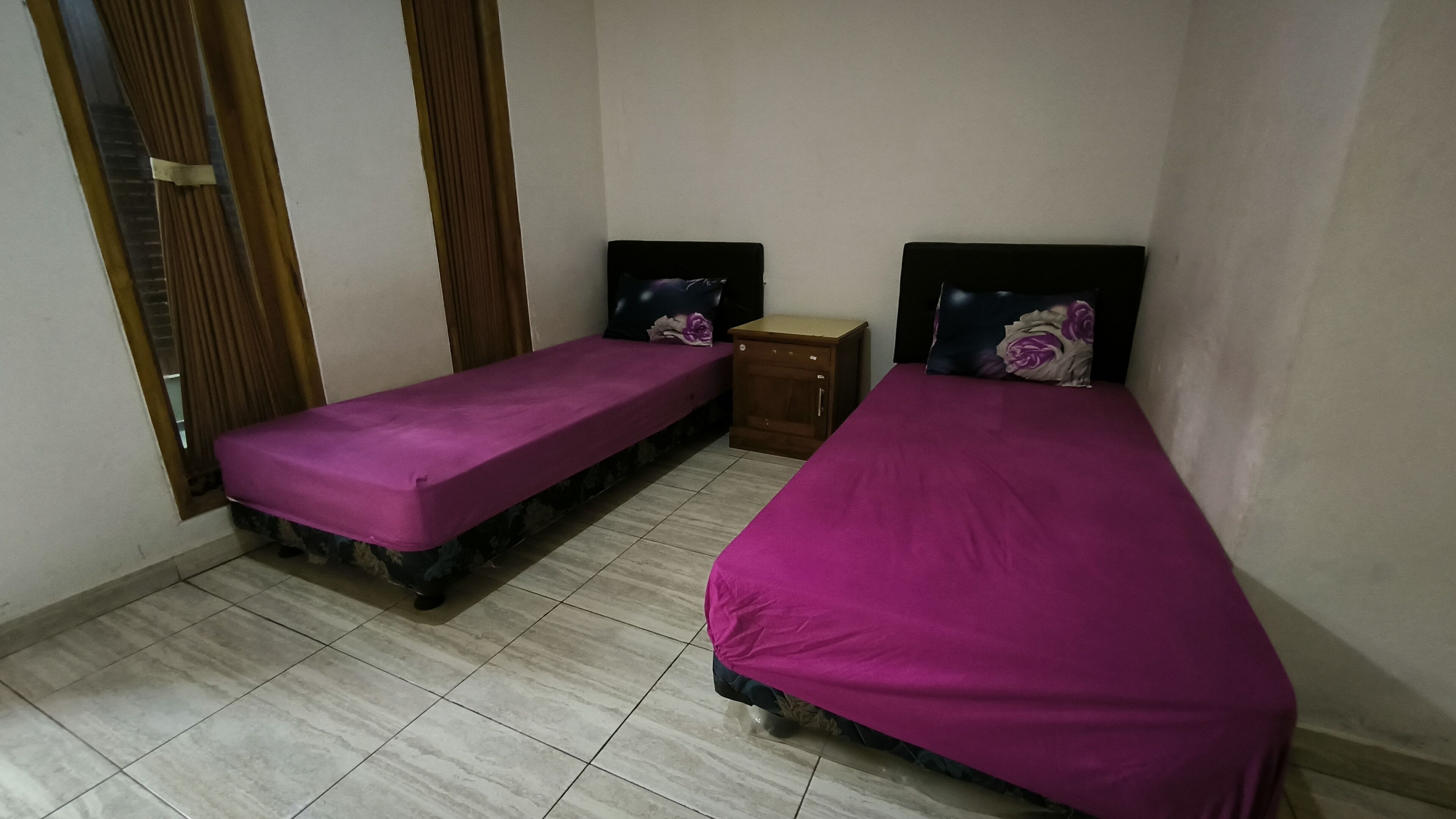 Una habitación pequeña y ordenada con dos camas individuales, una mesita de noche de madera y un ventanal con cortinas.