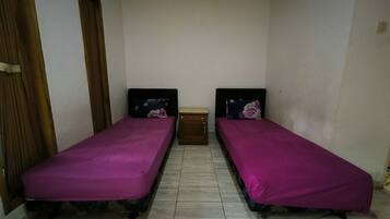 Une petite chambre avec deux lits simples, chacun recouvert d’une couvre-lit violette et agrémenté d’oreillers assortis. Une table de chevet en bois est placée entre les lits, et la chambre a des murs de couleur neutre et un sol carrelé.