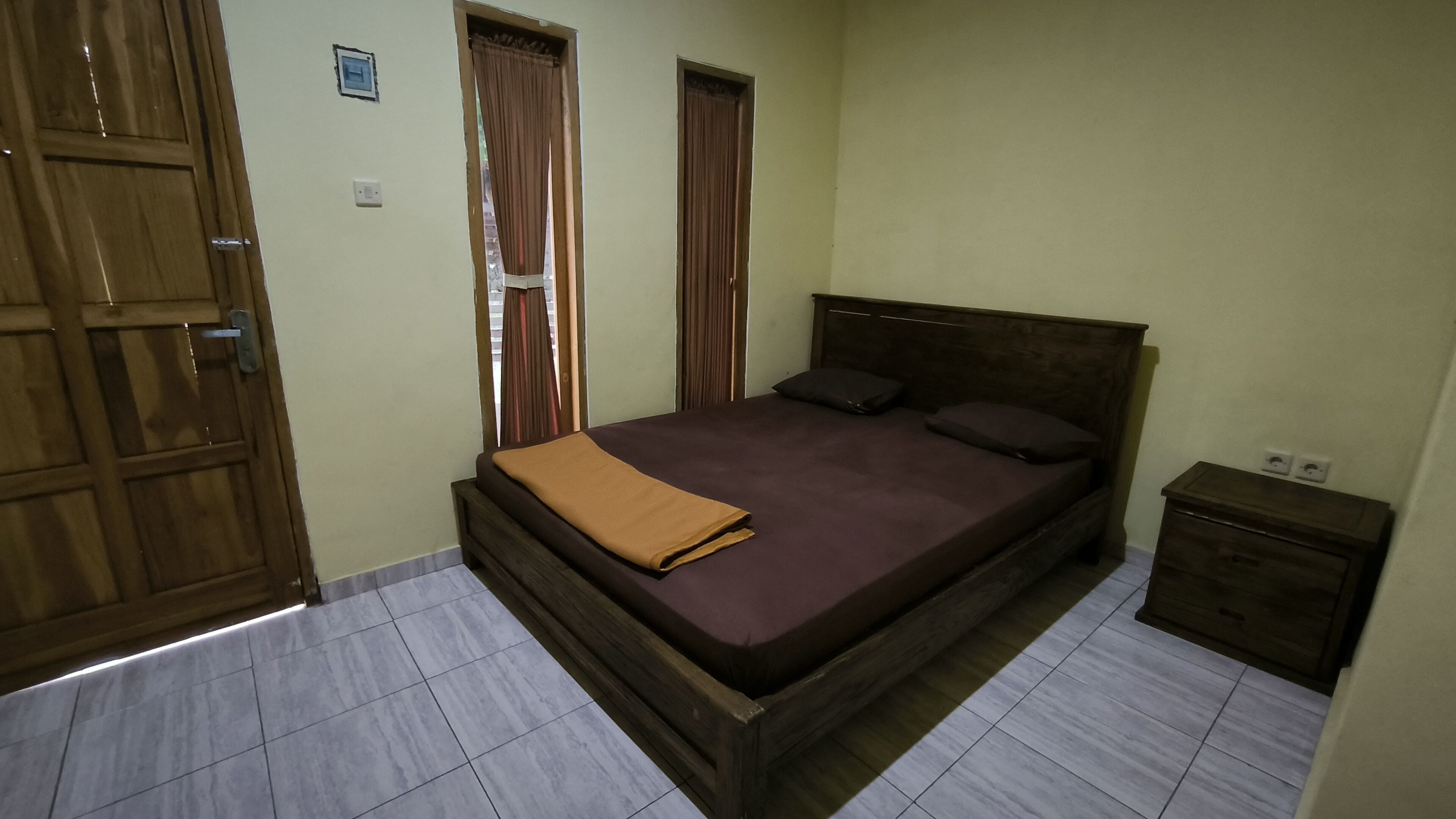 A bedroom with a wooden bed, two windows with curtains, a bedside table, and a tiled floor.