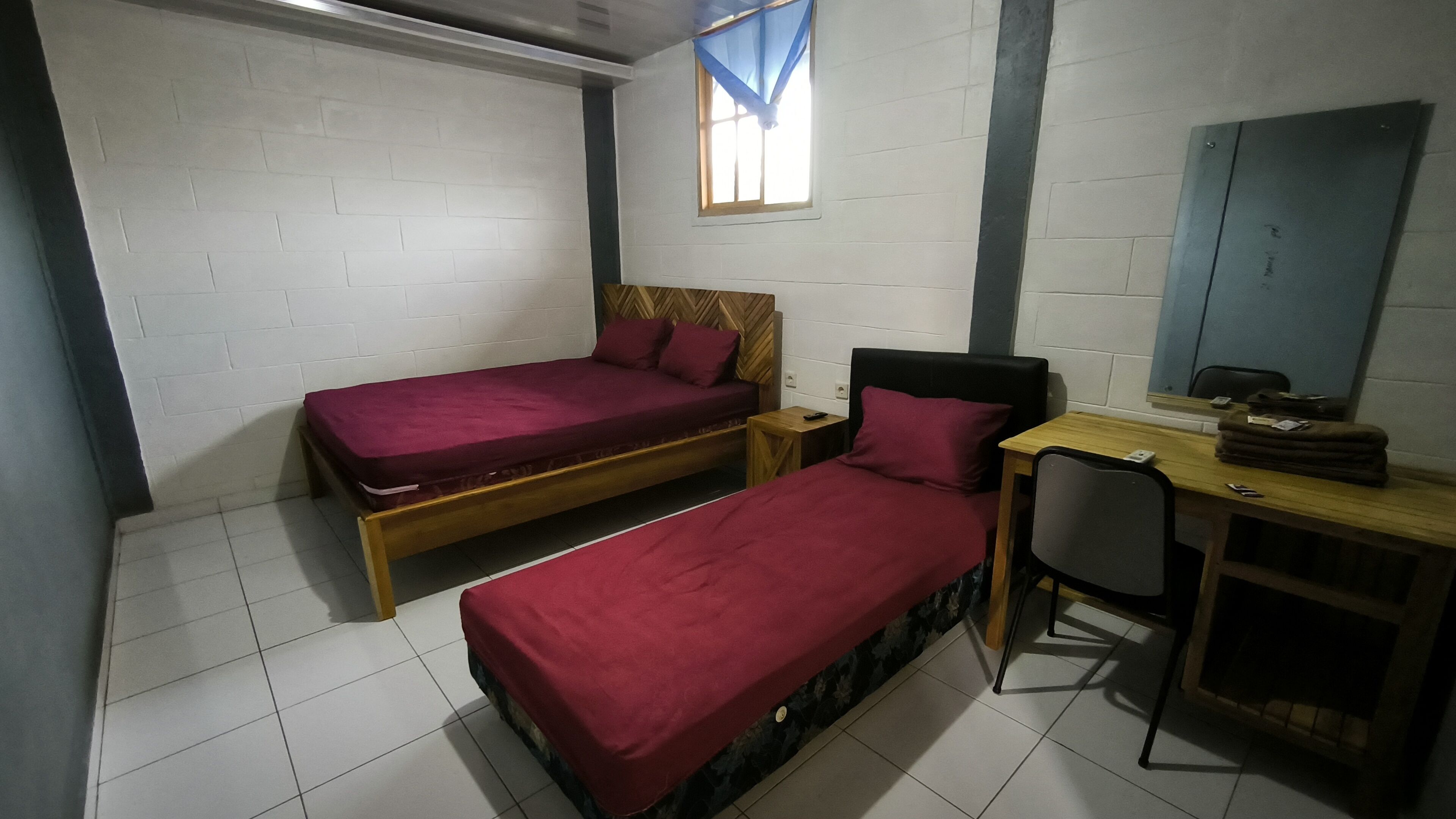 A dormitory room with two single beds, a desk, a chair, and a window with a curtain.