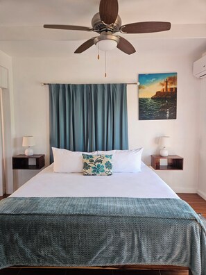 A bedroom with a bed, a ceiling fan, a painting, and two bedside tables with lamps. - Views from every angle - 2nd floor cozy villa (Port Antonio)