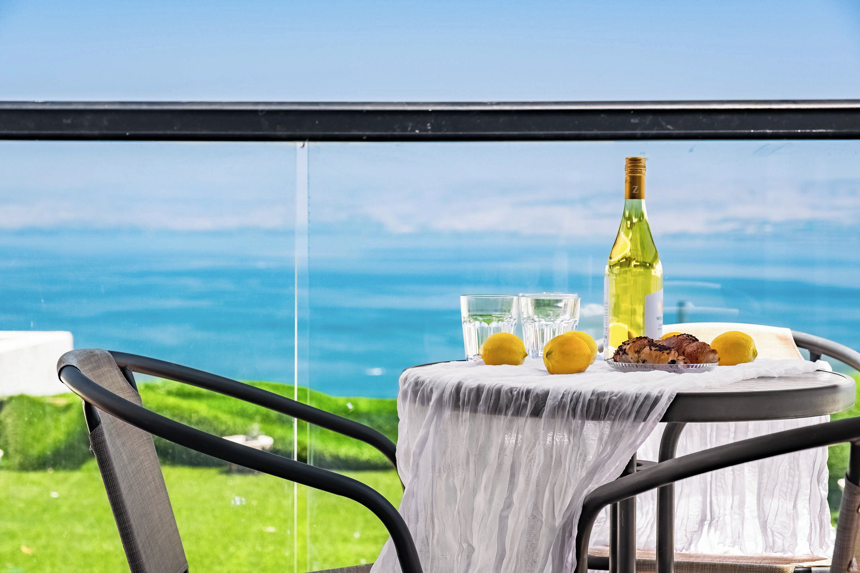 A table with a bottle, glasses, lemons, and food, overlooking a scenic view.