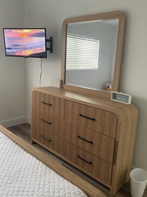 A bedroom with a wooden dresser, a television mounted on the wall, and a window with blinds.
