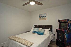 A bedroom with a bed, pillows, a ceiling fan, a beach-themed painting, and a Mortal Kombat arcade game. - Siesta Key Village • Pool • Walk to Beach • Pets (Sarasota)