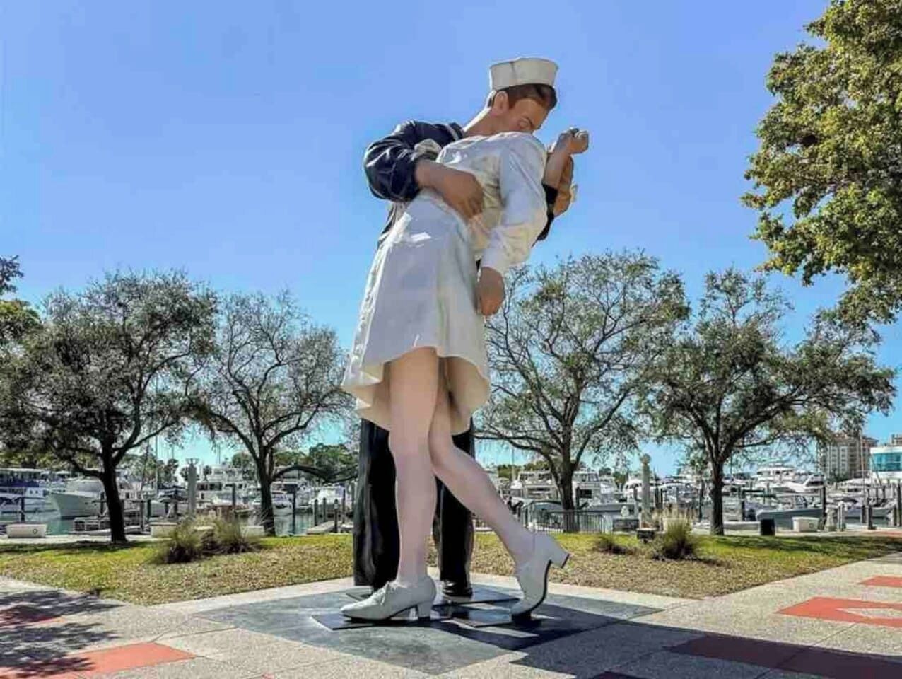 A statue of a man carrying a woman, set in an outdoor area with trees and a marina in the background.