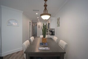 A dining area with a long table, white chairs, a hanging light fixture, and a plant centerpiece.