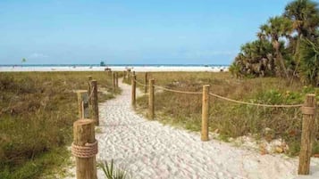 A sandy beach path with wooden posts and rope barriers, leading to a beach with palm trees and the ocean in the background.