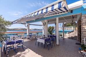Un área de comedor al aire libre con mesas y sillas, un toldo a rayas y vistas al mar y a las montañas.