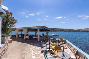 An outdoor dining area with stone flooring, tables, chairs, and a covered seating area overlooking a body of water.