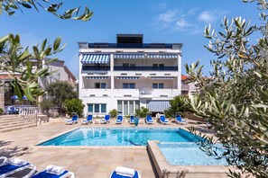 Un hotel de varios pisos con piscina, sillas de descanso azules y un cielo azul despejado.