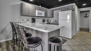 A modern kitchen with a marble island, bar stools, stainless steel trash can, and a refrigerator. - FLX Retreats Conesus Condos- APT B (Geneseo)