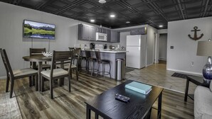A modern kitchen with grey cabinets, a white refrigerator, and a microwave. There is a dining area with a wooden table and chairs, and a TV mounted on the wall displaying a landscape. - FLX Retreats Conesus Condos- APT B (Geneseo)