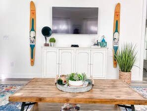 A room with a wooden table, a flat-screen TV mounted on the wall, and two surfboards on either side.