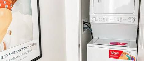 A compact laundry room with a washing machine and dryer, a framed picture on the wall, and a door leading to another room.