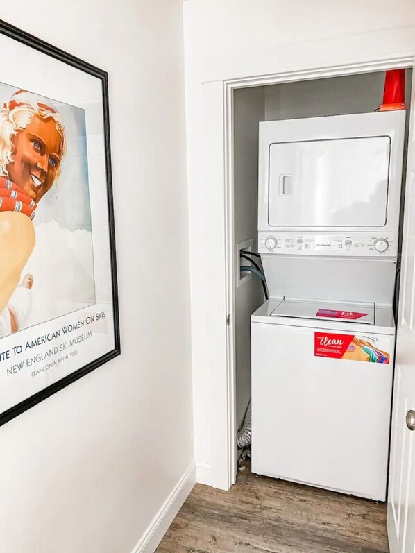 A compact laundry room with a washing machine and dryer, a framed picture on the wall, and a door leading to another room.