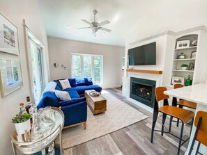 Un salon moderne avec un canapé bleu, une cheminée, un coin repas comprenant une table blanche et des chaises orange, et un grand téléviseur fixé au-dessus de la cheminée.