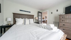 A hotel room with a large bed, a nightstand with a lamp, a dresser, and a television on the dresser. - New! Chatham Cottage- 1 Mile to Downtown & Beaches (Chatham)