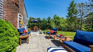 An outdoor patio with a brick wall, wooden furniture, and a blue umbrella. - New! Chatham Cottage- 1 Mile to Downtown & Beaches (Chatham)