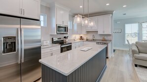 A modern kitchen with stainless steel appliances, a central island, pendant lighting, and a view to the living area. - Modern Little Beach House- Steps to the Beach (Gulf Shores)