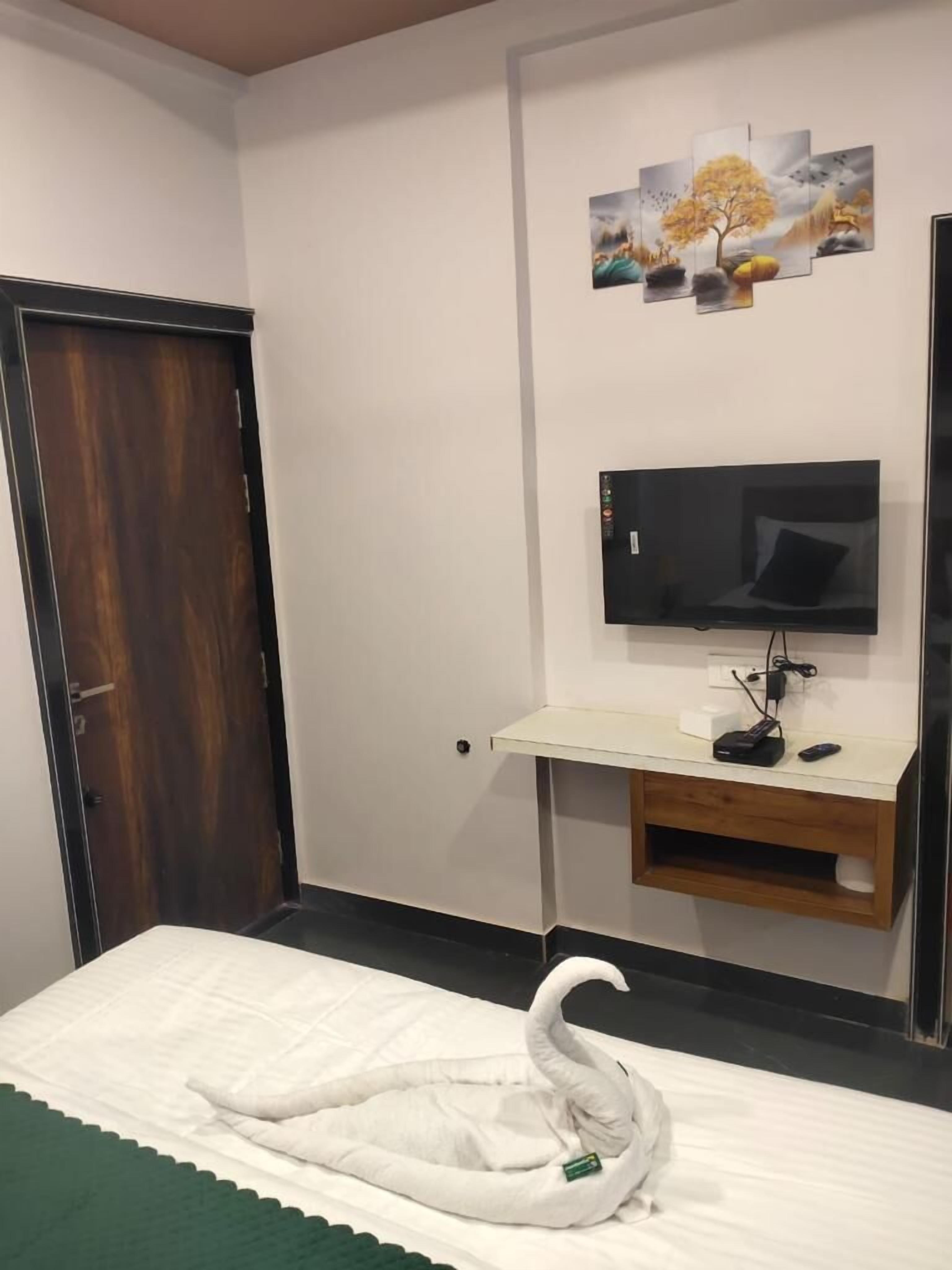 A hotel room with a bed, a TV mounted on a wooden stand, a painting on the wall, and a door.