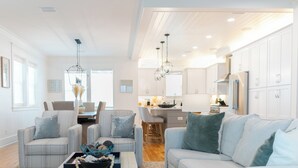 Amplia sala de estar con un sofá azul claro, dos sillones y una mesa de centro. Se ve una cocina al fondo con armarios blancos y electrodomésticos modernos.