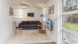 Ett modernt vardagsrum med trägolv, en grå soffa, ett litet bord med en lampa och ett stort fönster med utsikt över träd.