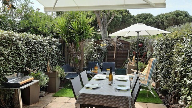 An outdoor patio with a table set for dining, surrounded by lush greenery and a white umbrella.