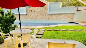 Une terrasse avec une table ronde et des chaises, un parasol rouge et une piscine en arrière-plan.