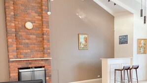 A living room with a brick fireplace, a black sofa, a wooden coffee table, and a kitchen area with bar stools. - The peaceful retreat in a tranquil court (Mulgrave)