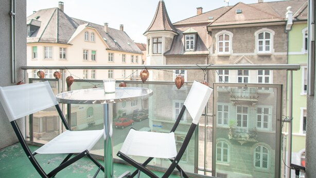 A balcony with two white chairs and a small round table, overlooking a row of traditional European buildings.