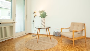 A room with a wooden floor, a round rug, a chair, a table with a vase of flowers, and a radiator.