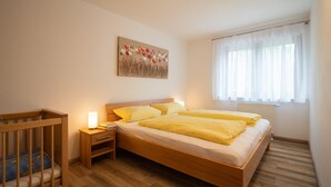 A bedroom with a wooden bed, a nightstand, a crib, and a window with sheer curtains. - Ferienwohnung Simssee 60 qm (Rimsting)