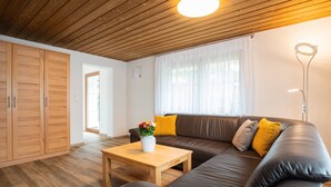 A modern living room with a wooden ceiling, a brown leather sofa, a wooden coffee table with a vase of flowers, and a floor lamp. - Ferienwohnung Simssee 60 qm (Rimsting)