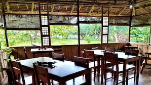 A restaurant with wooden tables and chairs, large windows, and a view of greenery outside. - The Lazy Tiki (Buenavista)