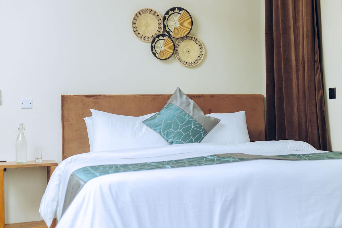 A neatly made bed with white bedding and a green and white patterned throw. There are two pillows, one with a geometric pattern and another plain white. A wooden nightstand with a glass bottle and a small glass is to the left.