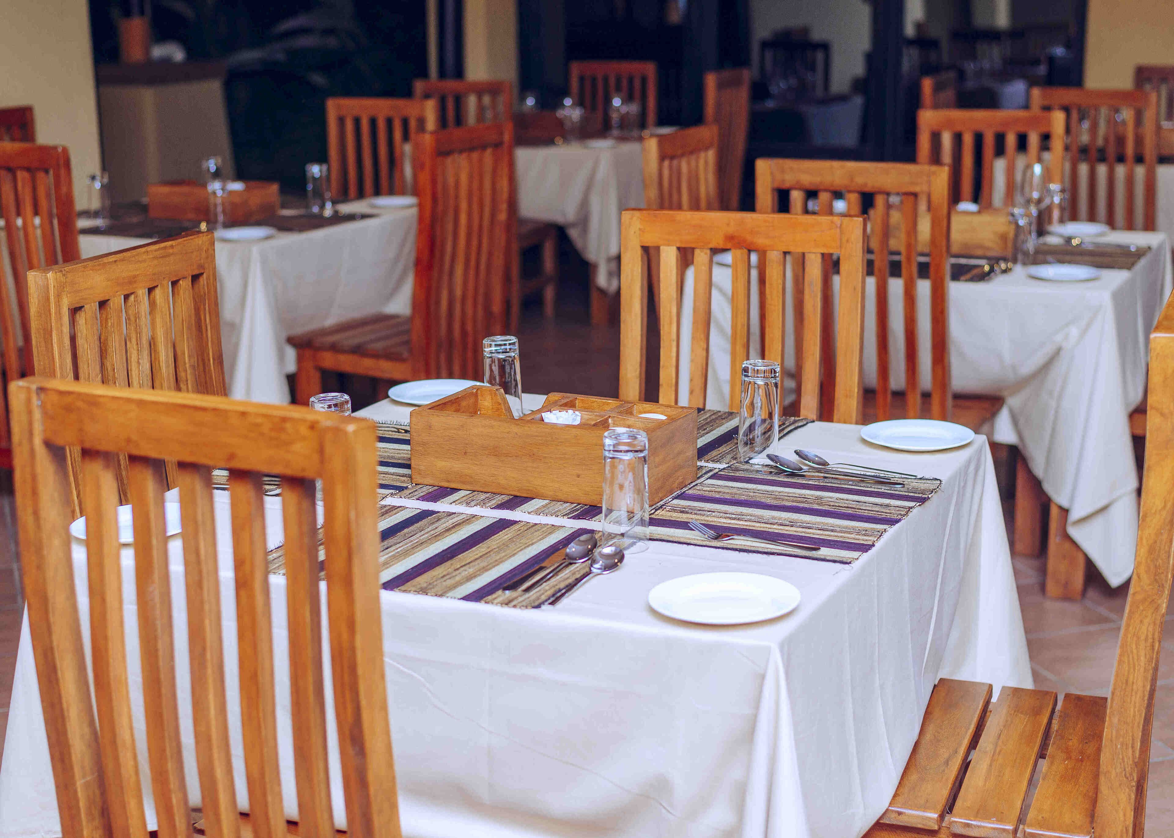 A dining area with wooden chairs and tables, each set with white tablecloths, plates, and cutlery.