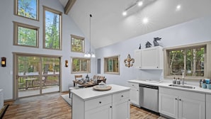 A modern kitchen with white cabinetry, a large island, and a dining area with a view of the outdoors. - Hot Tub Chalet, Fire Pit, Lake Access, Dogs OK (Blakeslee)