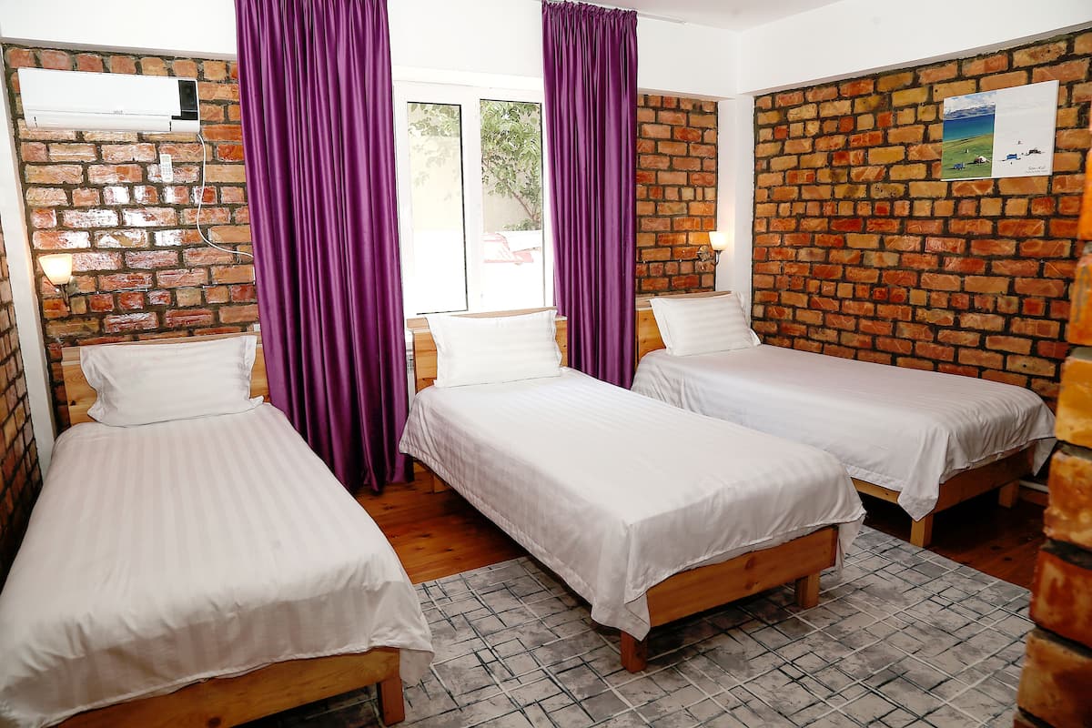 Une chambre avec trois lits simples, des rideaux violets, un mur en briques et un appareil de climatisation.