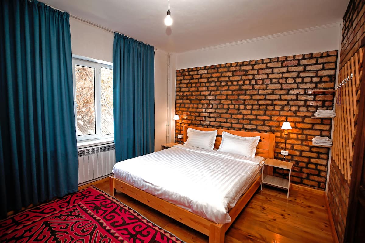Une chambre avec un mur décoré d’un motif en briques, un lit en bois, un tapis rouge à motifs et une fenêtre avec des rideaux bleus.