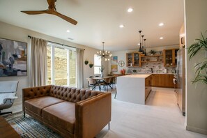 A modern living room with a brown leather sofa, a dining area with a table and chairs, a kitchen with wooden cabinets and a stone backsplash, and a ceiling fan. - The Q by Velo - Stylish Mid-Century Modern Home (Albuquerque)