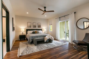 A bedroom with a bed, bedside tables, a ceiling fan, a large window with curtains, and a view of the outdoors. - The Q by Velo - Stylish Mid-Century Modern Home (Albuquerque)