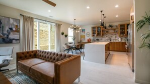 Un salon moderne avec un canapé en cuir marron, un coin repas avec une table et des chaises, une cuisine avec des meubles en bois et un pare-feu en pierre, et un ventilateur de plafond.