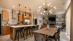 Une cuisine moderne avec des meubles en bois, un pare-feu en marbre et un îlot central doté de tabourets de bar. On y trouve également un coin repas avec une table et des chaises en bois, et un grand lustre au-dessus.